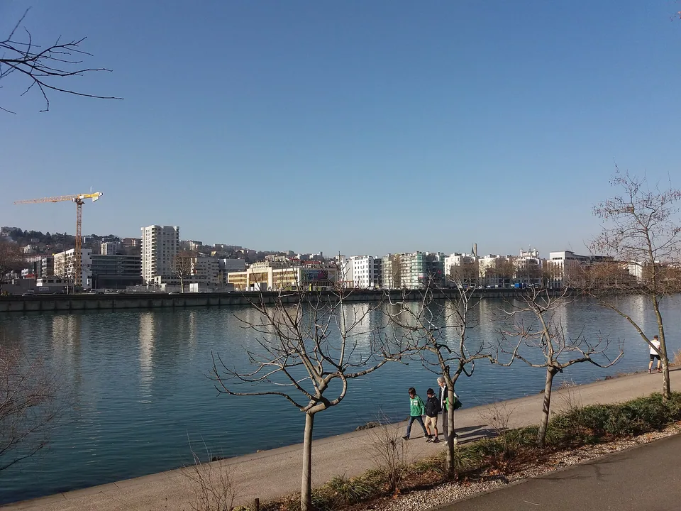 河对岸 Confluence 的不同年代建筑和施工塔吊并置，近景为河滨步道上的行人。