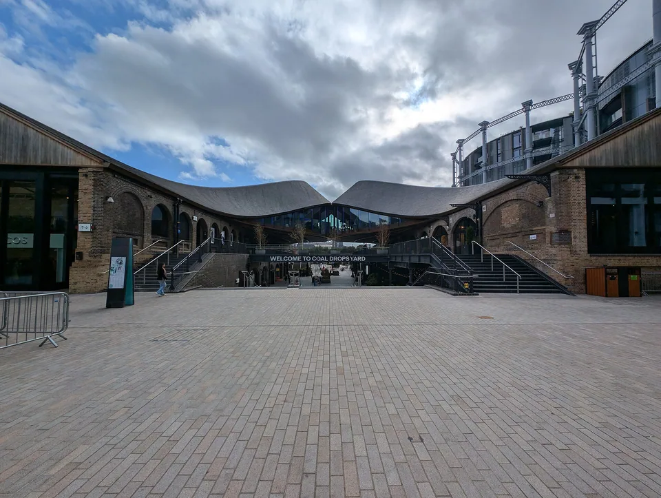 从北侧望向 Coal Drops Yard，两侧砖砌建筑、弧形屋顶、楼梯与中部步行通道。