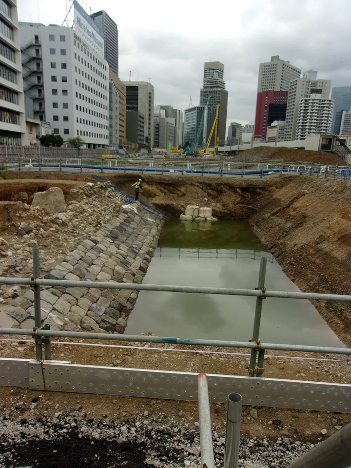 高轮筑堤石墙近景，旁边为开挖沟槽和施工设施，远处可见城市建筑。